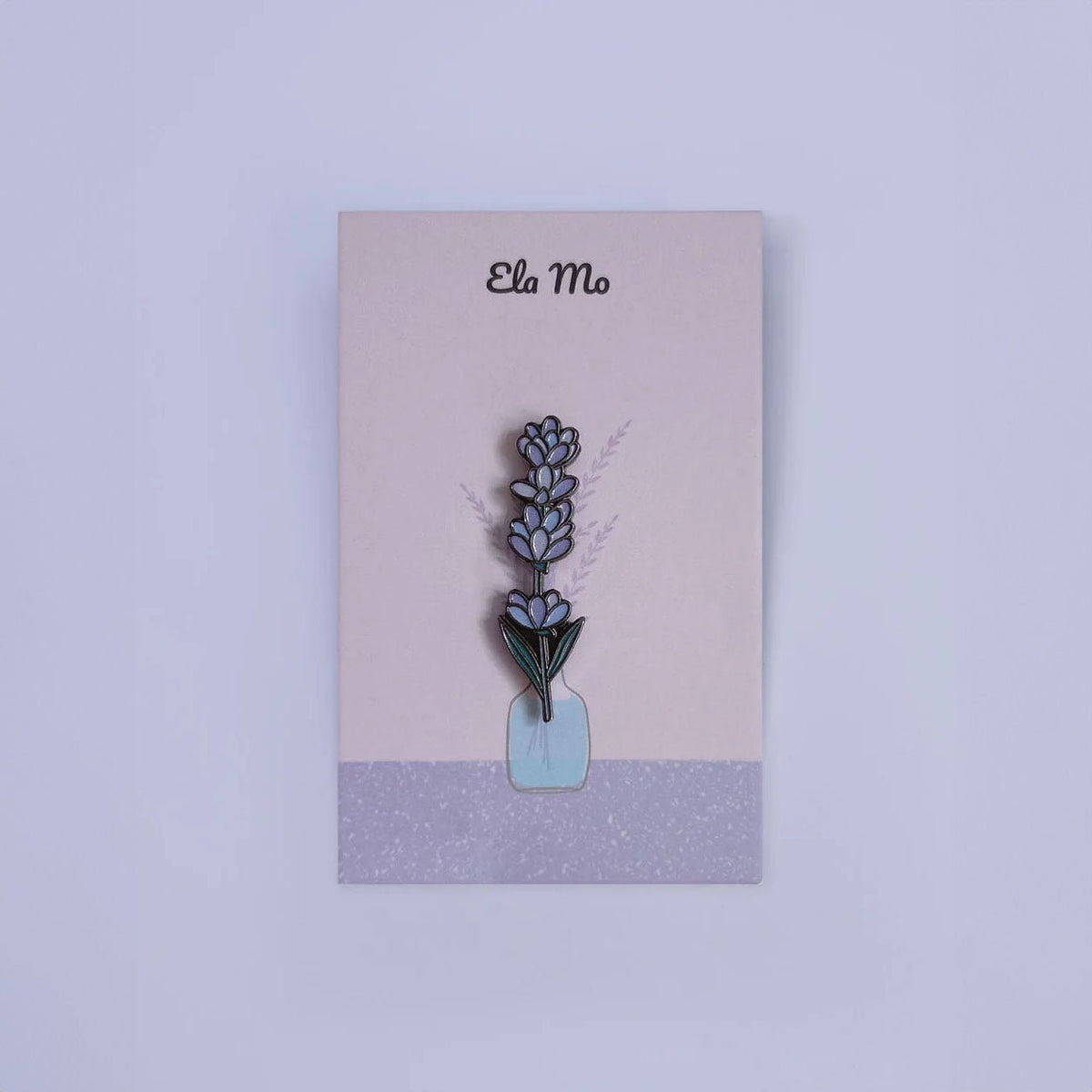 Ela Mo™ Mini Pin für Rucksäcke | Lavender. Ein kleiner Lavendel-Pin auf einer Karte befestigt, vor lila Hintergrund.