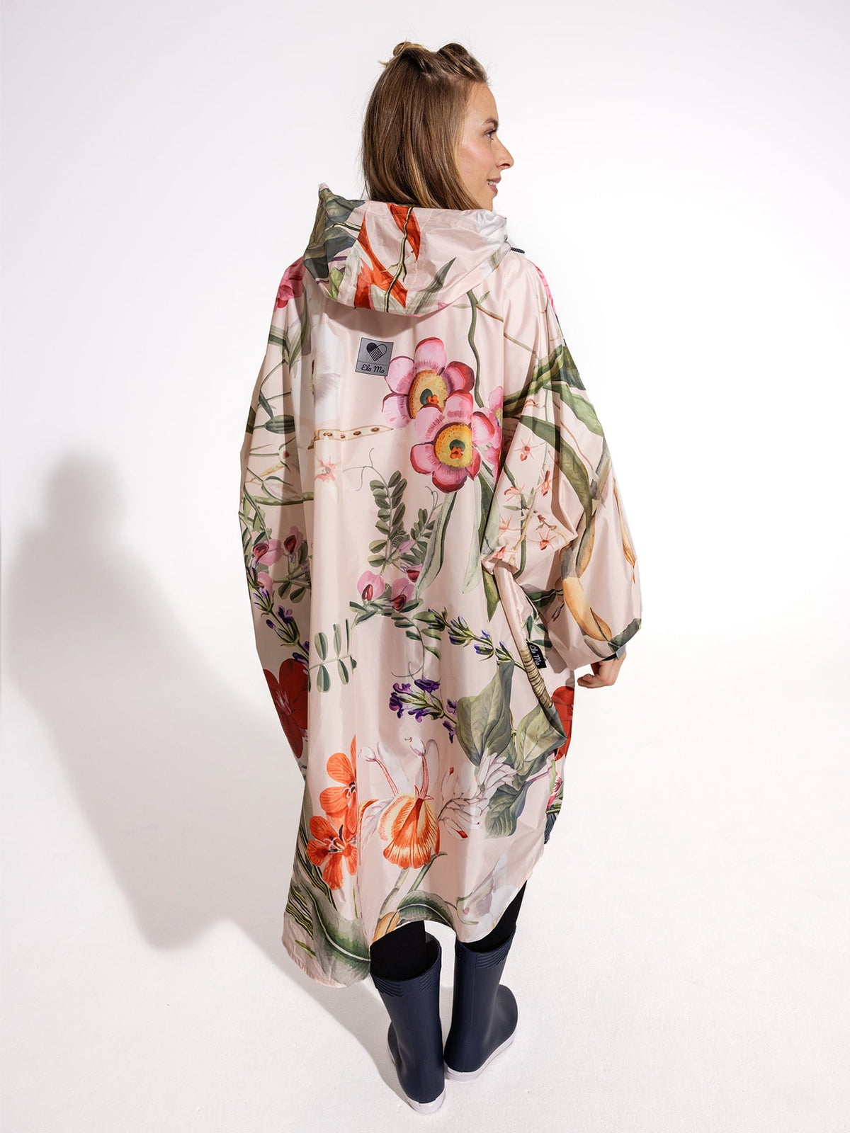 Ela Mo™ Regenponcho | Luxe. Eine Frau im Regenponcho mit Kapuze, Ansicht von hinten.