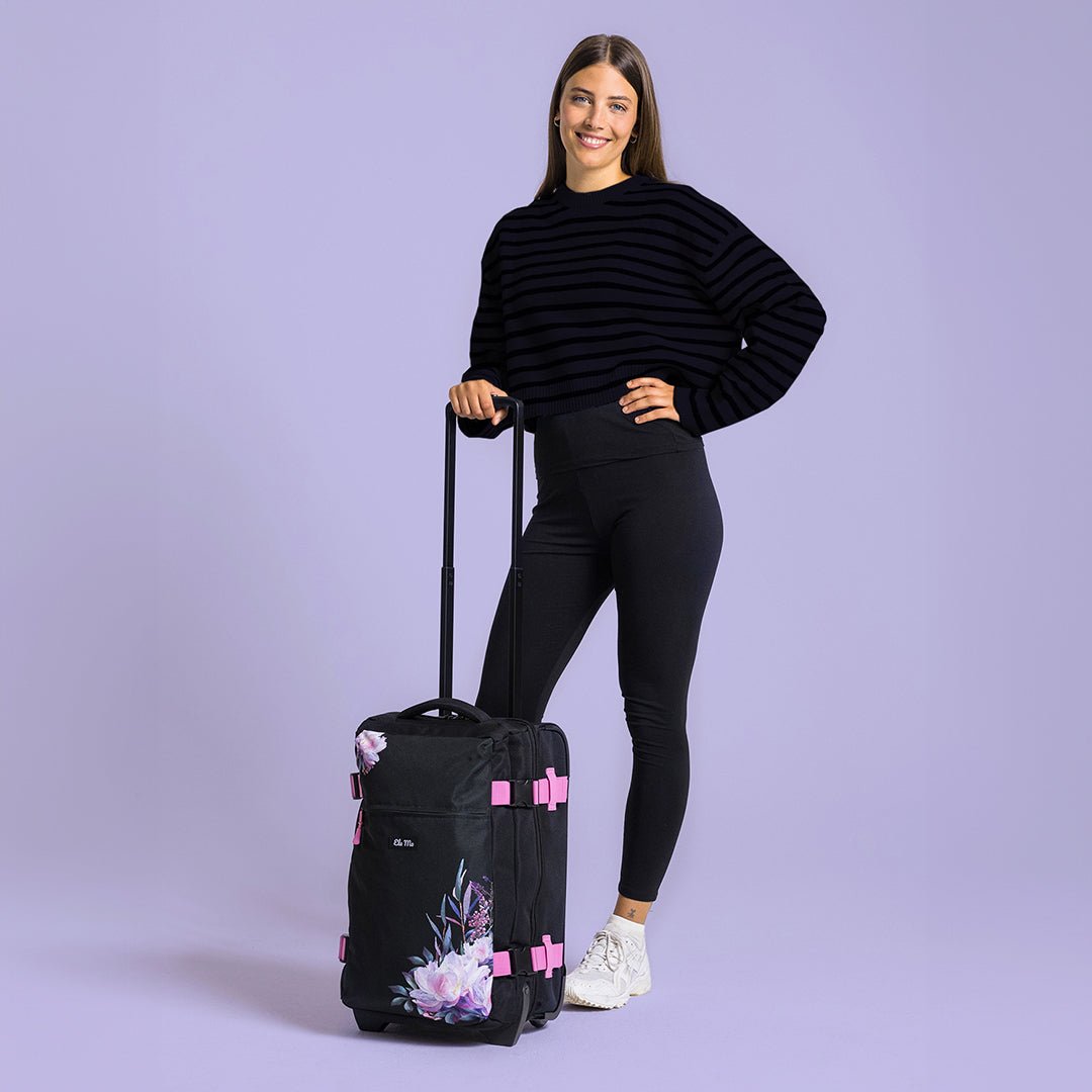 Ela Mo™ Reisetasche mit Rollen 42 Liter | Beauty in Darkness. Eine Frau neben einer Reisetasche mit hochgezogenem Griff.