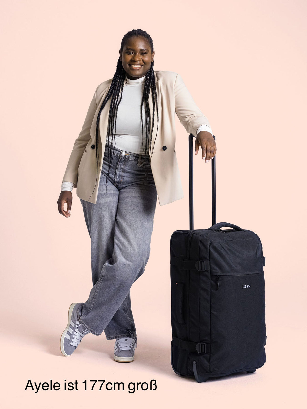 Ela Mo™ Reisetasche mit Rollen 78 Liter | Black. Eine Frau neben einer Reisetasche mit hochgezogenem Griff.