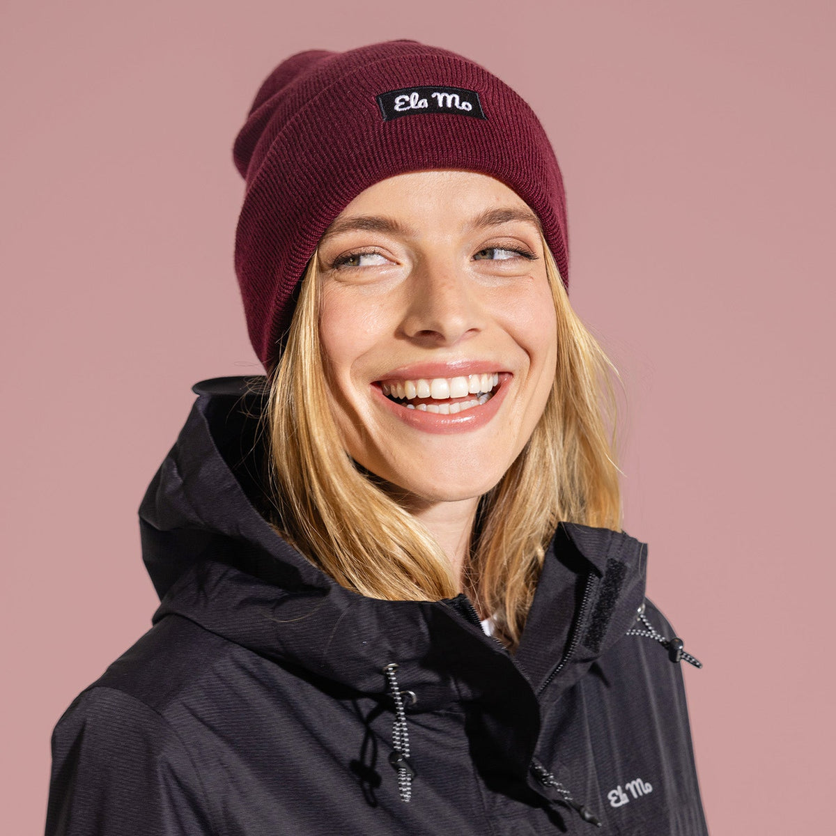 Ela Mo™ Beanie | Burgund. Eine Frau mit einer warmen Mütze in Burgunderrot und Herstellerlogo darauf.