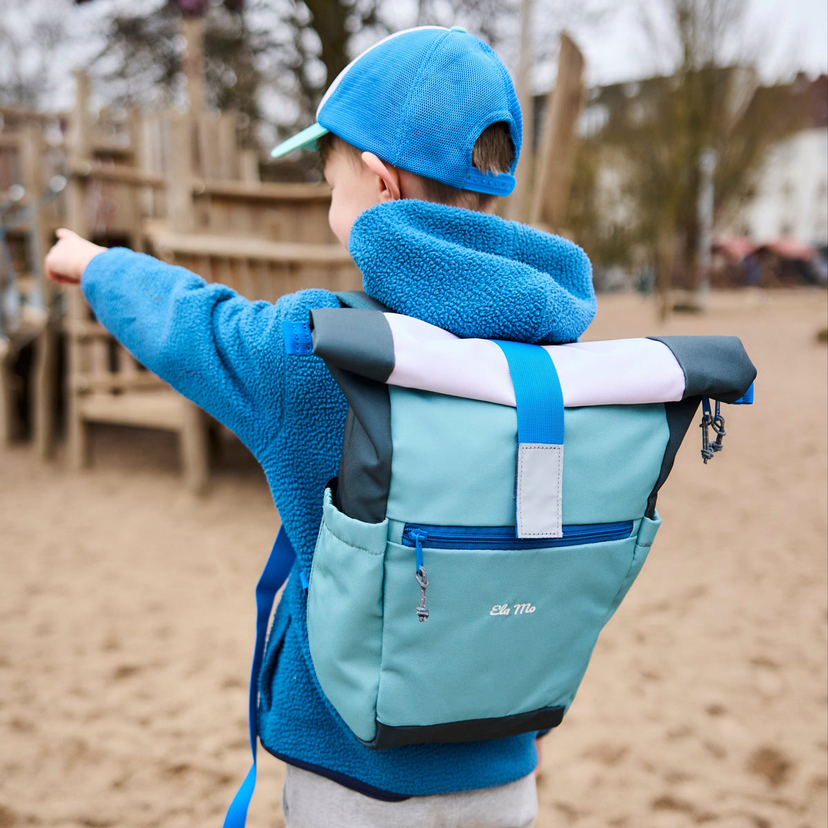 Ela Mo™ Kinderrucksack (6-10 Jahre) | Blue Mint. Ein Junge auf dem Spielplatz, der einen blauen Rucksack trägt.