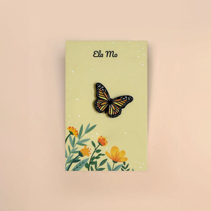 Ela Mo™ Mini Pin für Rucksäcke | Butterfly. Ein kleiner Schmetterlings-Pin auf einer Karte befestigt, vor gelbem Hintergrund.