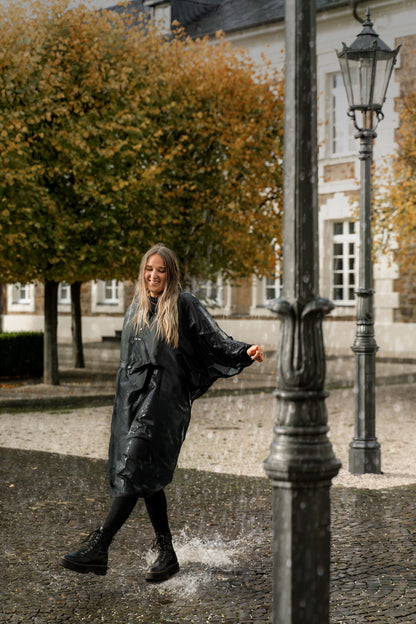 Ela Mo™ Regenponcho | Black. Frau mit Regenponcho an einem regnerischen Tag auf der Straße.